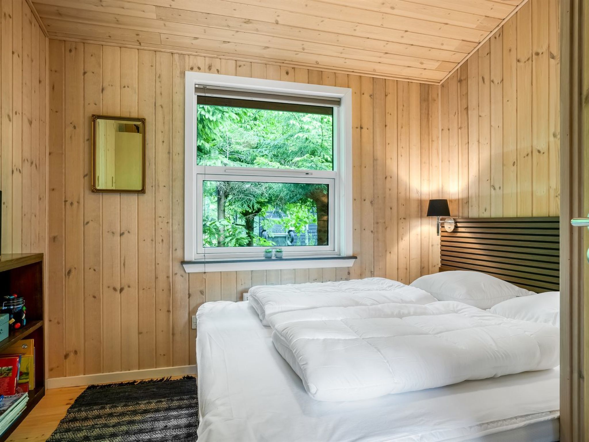 A bedroom with wooden walls, a large window with a view of greenery, a bed with white bedding, and a bedside table with a lamp.