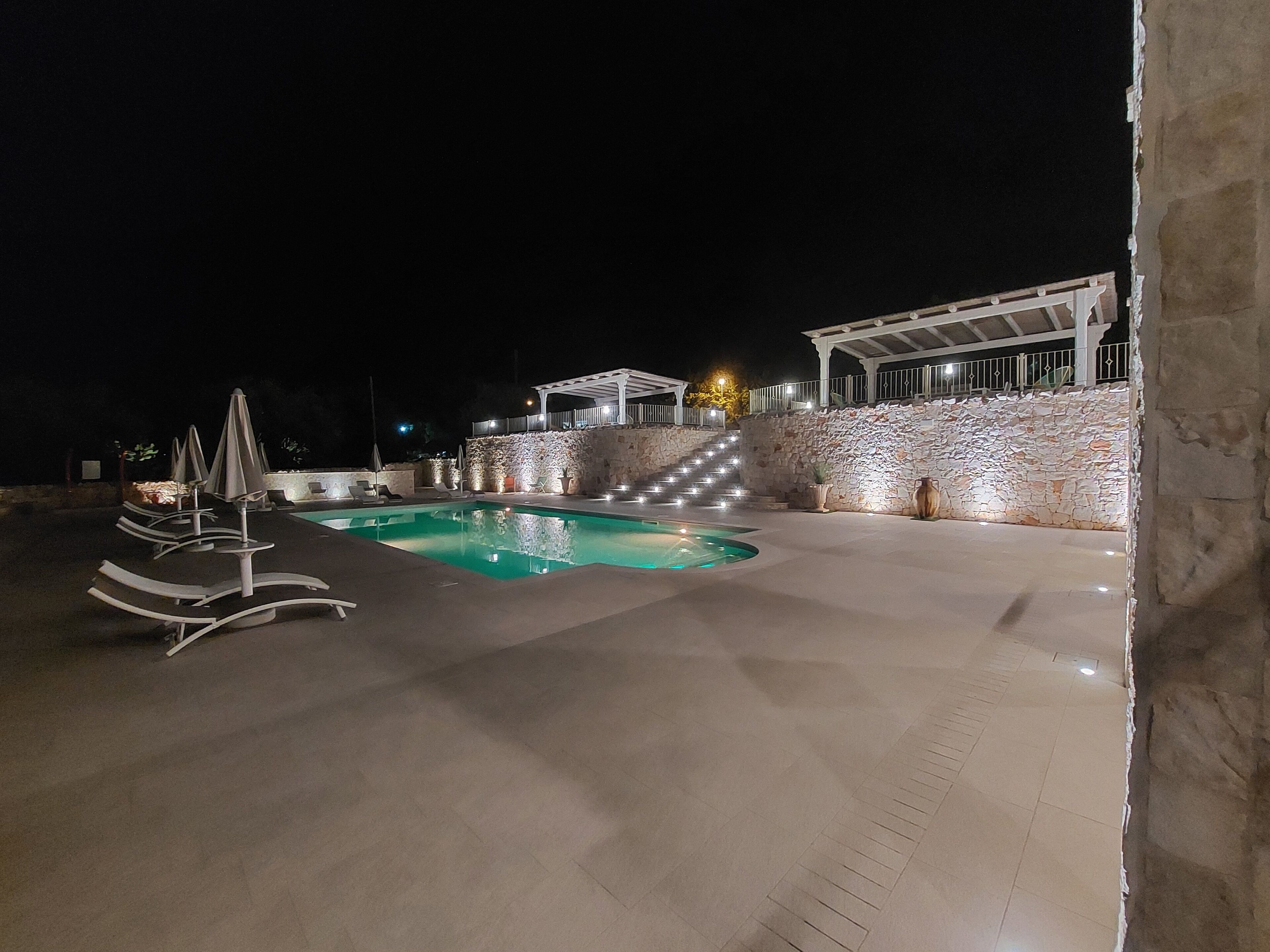 Piscina com espreguiçadeiras e guarda-sóis, uma parede de pedra e uma escadaria que leva a uma área coberta.