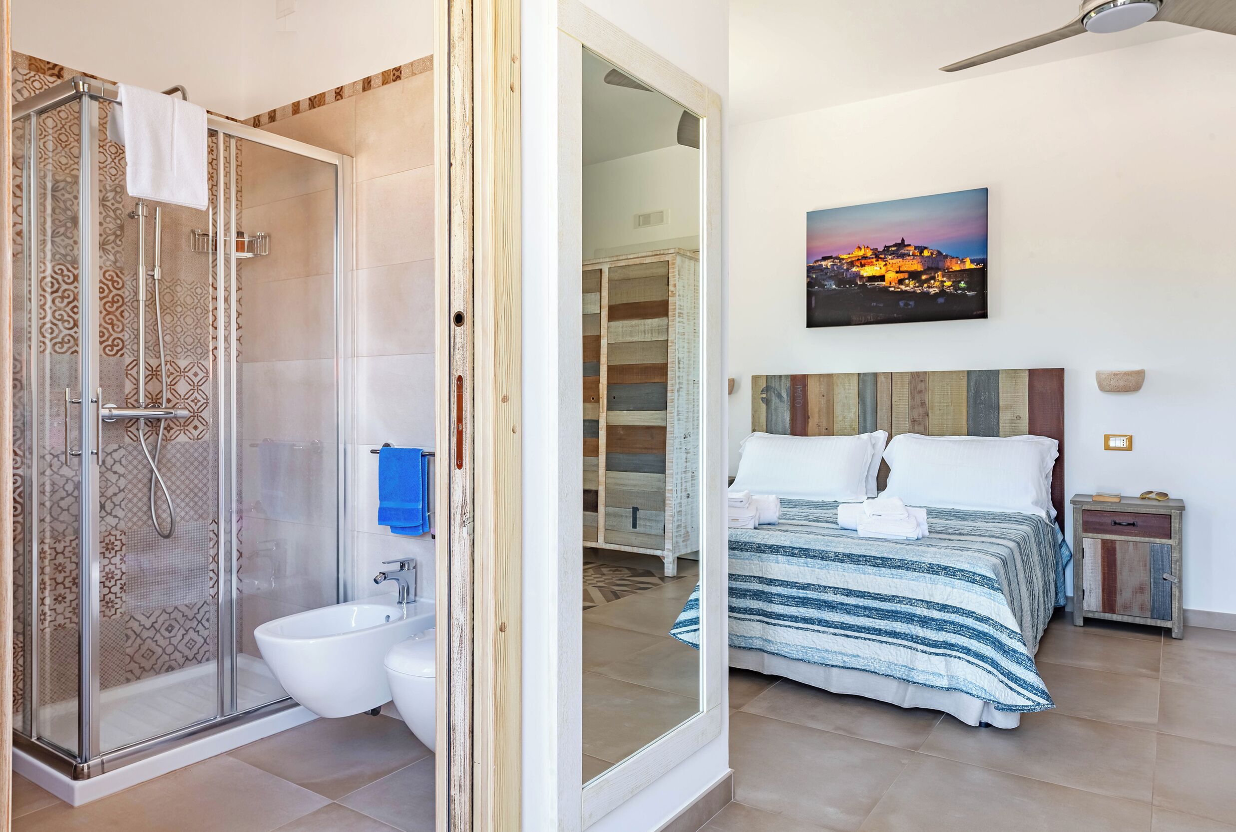 Casa de banho com cabine de duche em vidro, sanita e lavatório. Há uma cama com colcha e almofadas riscadas, uma mesa de cabeceira com candeeiro, e um quadro com um pôr do sol na parede.