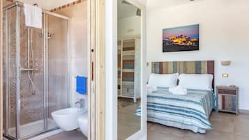 Casa de banho com cabine de duche em vidro, sanita e lavatório. Há uma cama com colcha e almofadas riscadas, uma mesa de cabeceira com candeeiro, e um quadro com um pôr do sol na parede.