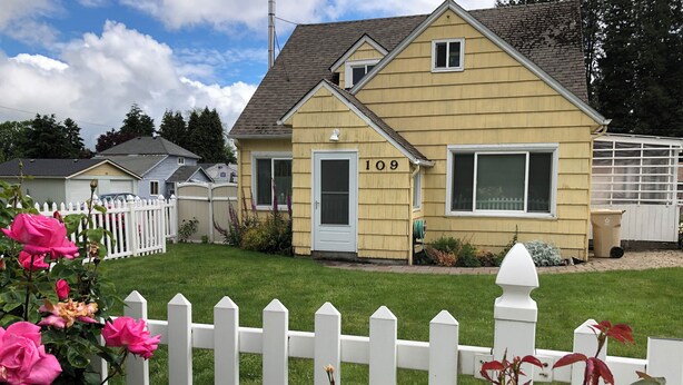Une maison jaune, portant le numéro 109, une clôture blanche en bois et des rosiers roses au premier plan.