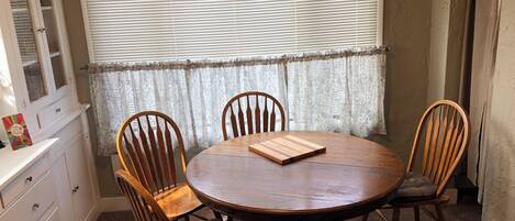 Un comedor con una mesa redonda de madera, cuatro sillas de madera, una cortina de encaje y una ventana con persianas.