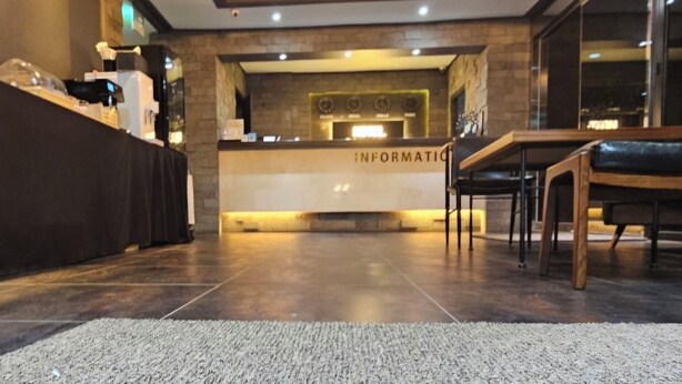 Un área de recepción con un mostrador identificado como 'INFORMACIÓN', una zona de estar con una mesa y sillas, y un reloj en la pared.