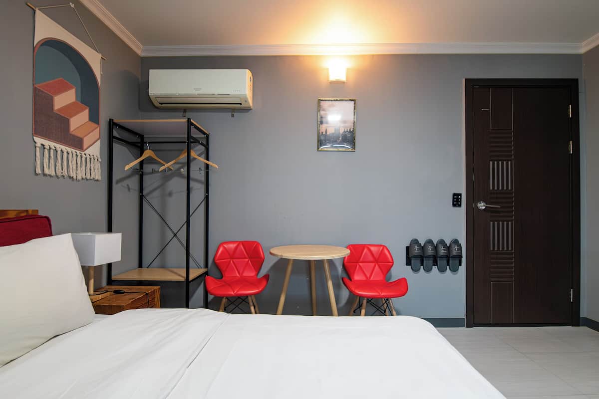 Une chambre d’hôtel comprenant un lit, une table avec deux chaises rouges, un porte-manteau, un climatiseur, un tableau encadré et une porte avec un espace pour ranger les chaussures.