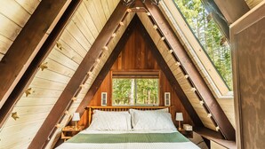 Une chambre confortable située sous les toits, avec un plafond en pente, des poutres apparentes, une grande fenêtre et un lit recouvert d’une literie verte.