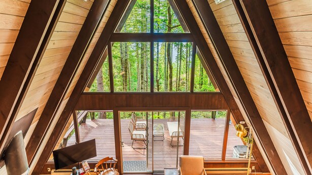 Une cabane en bois avec un toit triangulaire, de grandes fenêtres et une terrasse dotée de meubles d’extérieur.