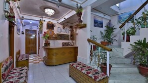 Un vestíbulo de hotel con suelo a cuadros, bancos de madera, un mostrador de recepción y una escalera adornada con plantas en macetas.