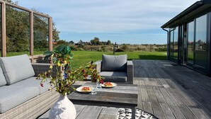 An outdoor deck with a sofa, a dining table with plates and glasses, and a large window overlooking a grassy area.