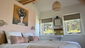 A bedroom with a bed, two bedside lamps, a painting on the wall, a window with a view of trees, and a hanging light fixture.