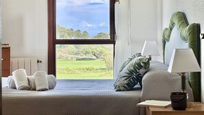 Une chambre avec une grande fenêtre, un lit avec des oreillers, une table de chevet avec une lampe, et une vue sur un paysage verdoyant.
