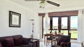 Un salon avec un canapé, une table basse et un fauteuil. On y trouve également une table à manger avec des chaises, ainsi qu’une fenêtre avec des rideaux.