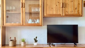 Un meuble en bois avec des portes en verre contenant des ustensiles de cuisine, un téléviseur posé sur un meuble en bois, et deux plantes en pot.