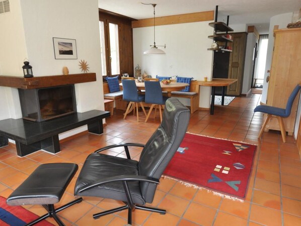 A living room with a fireplace, a dining area with a table and chairs, and a kitchen area with cabinets and a hanging light fixture.