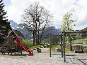 Un terrain de jeux avec un toboggan rouge et des balançoires, le tout situé dans un cadre magnifique, avec des montagnes et des arbres en arrière-plan.