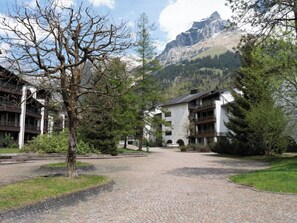 Une cour avec un chemin pavé menant à un bâtiment doté de nombreux balcons, entouré d’arbres et avec une chaîne de montagnes en toile de fond.