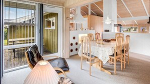Un vaste espace de salle à manger avec une table et des chaises en bois, un balcon offrant une vue agréable et une cuisine moderne dotée de meubles blancs et d’un éclairage suspendu.
