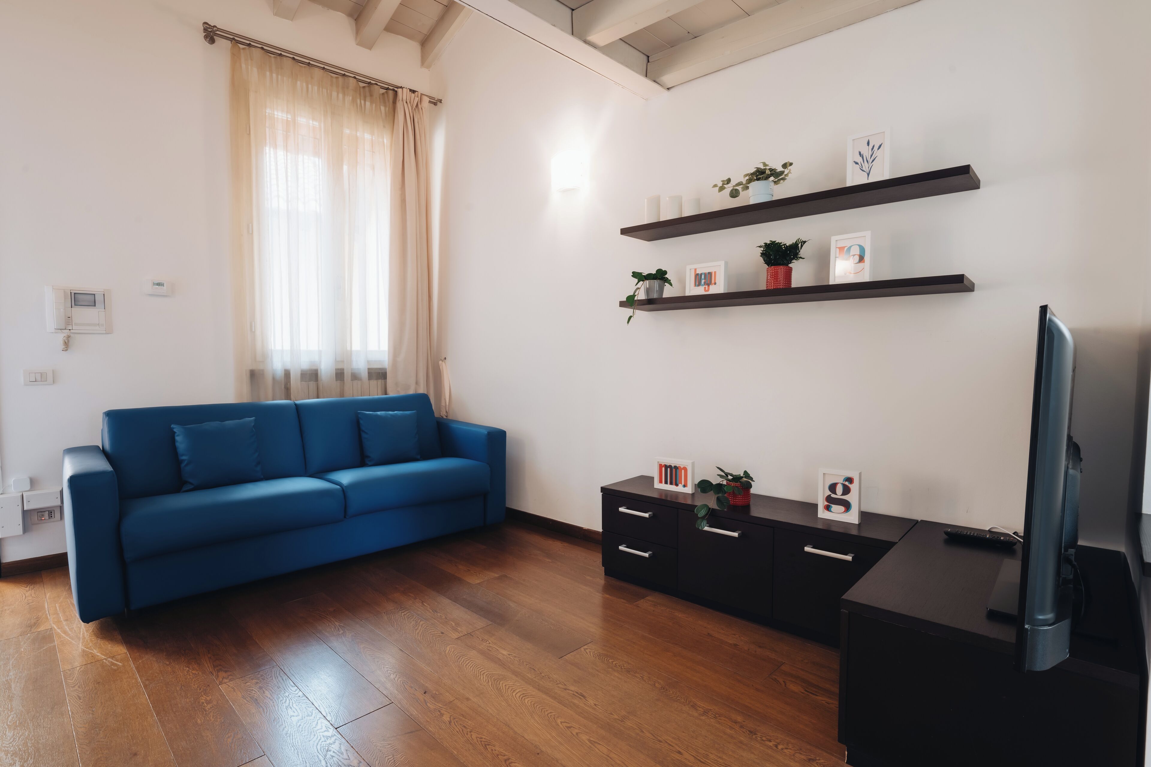 Un salon moderne avec un canapé bleu, un plancher en bois, un meuble TV noir et des étagères murales ornées d’objets décoratifs.