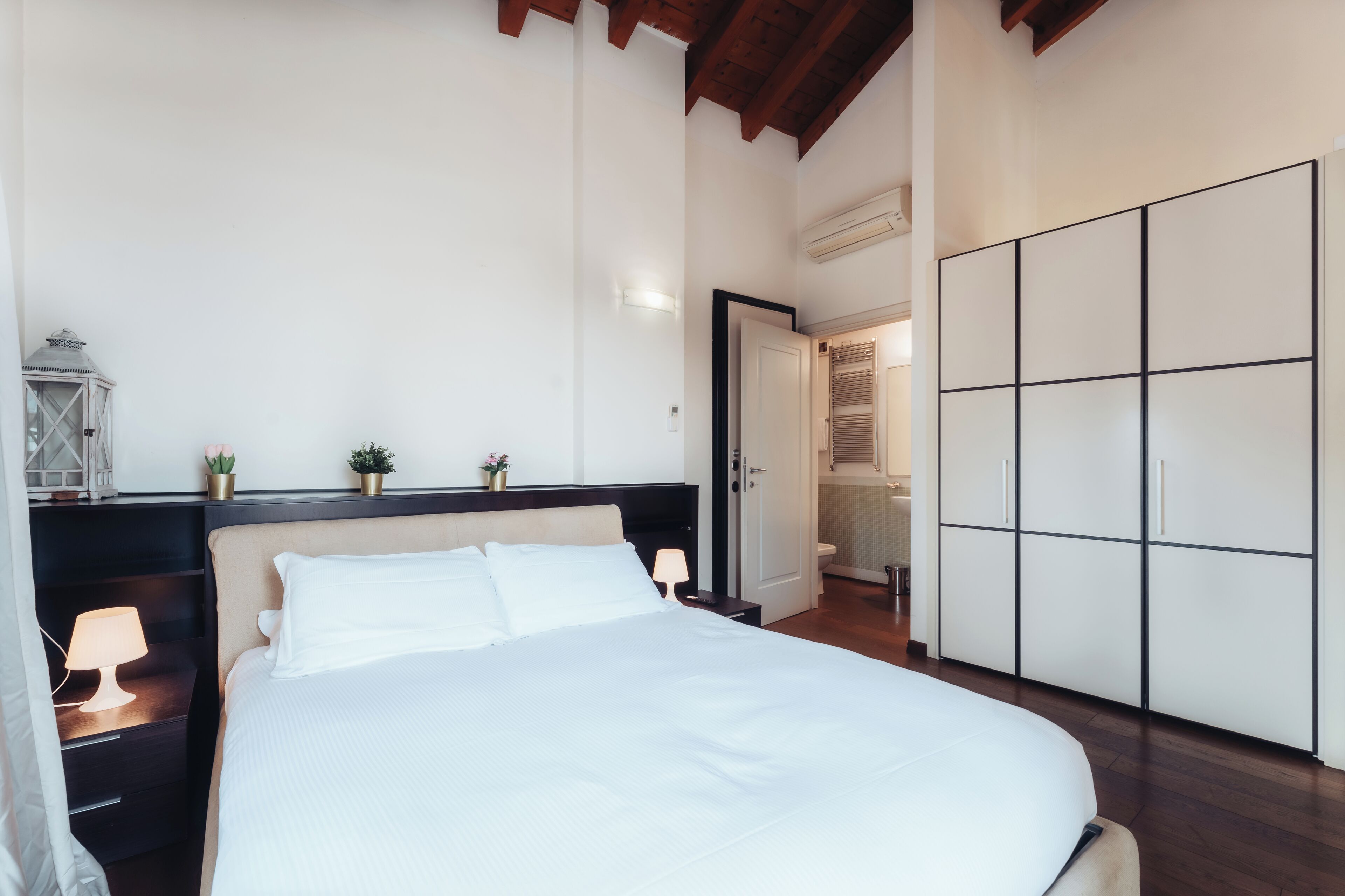 A bedroom with a large bed, a nightstand with a lamp, a wardrobe, and a door leading to a bathroom.