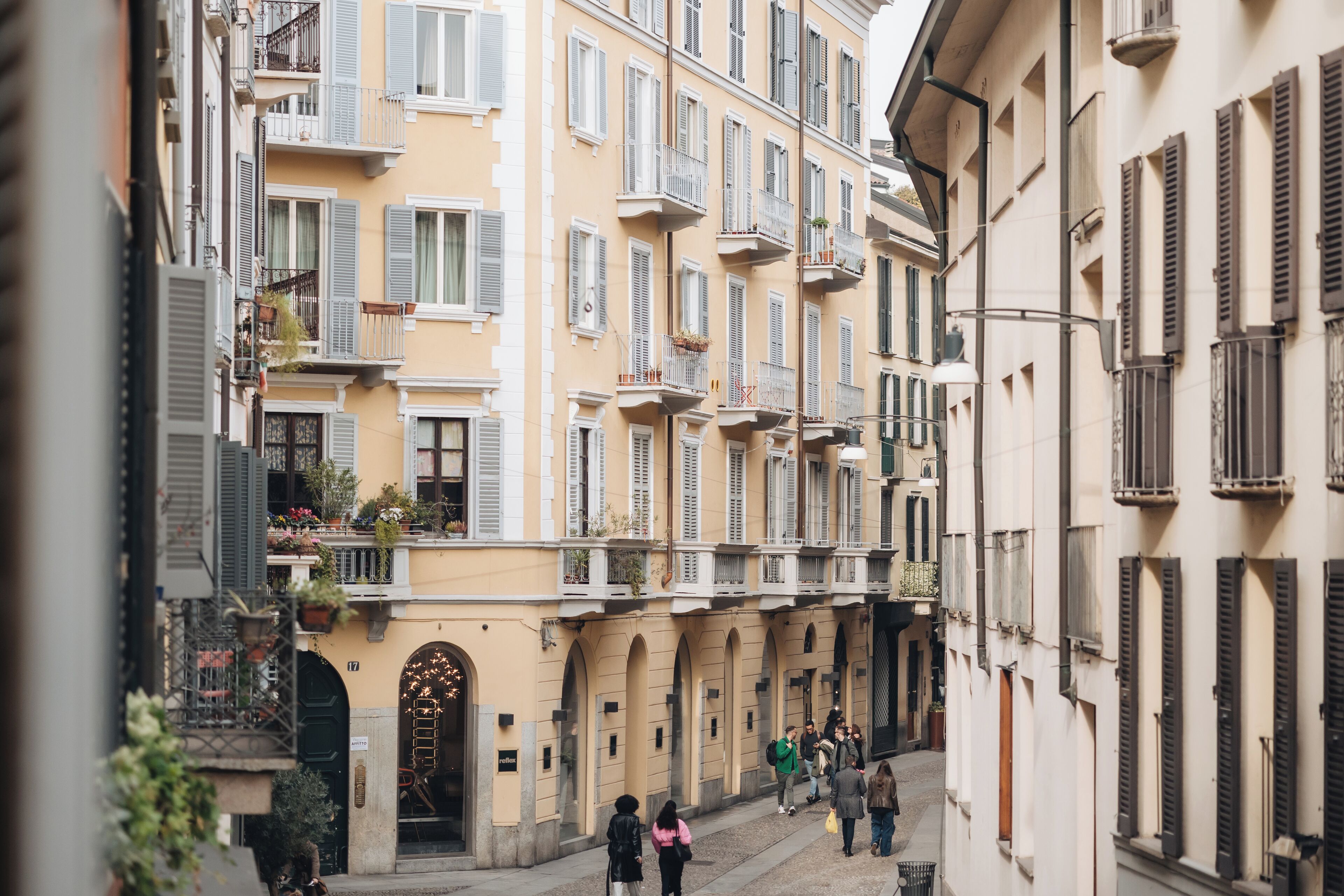 Une rue étroite bordée d’immeubles à plusieurs étages, de balcons ornés de plantes et où l’on croise quelques piétons.