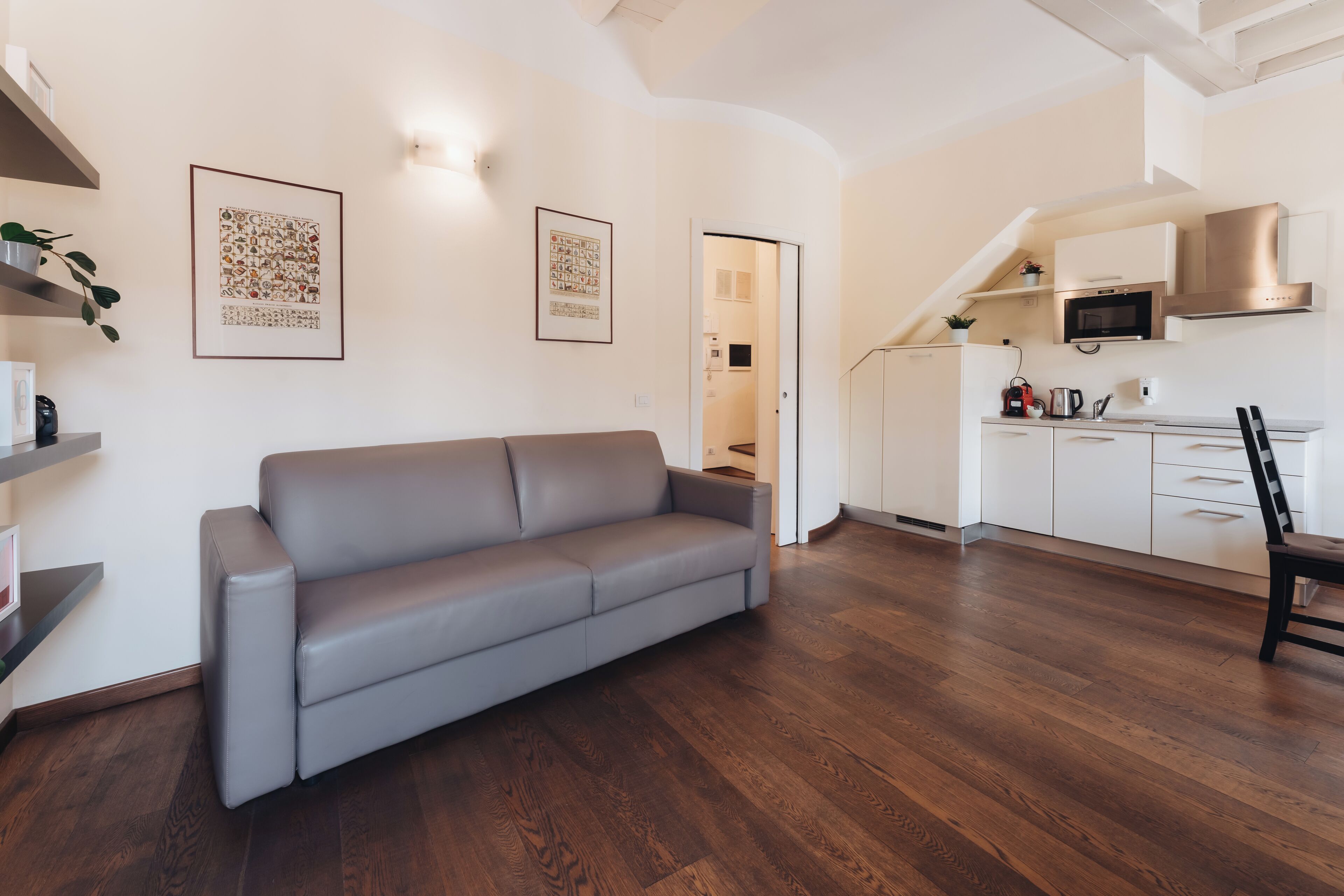 A modern living room with a grey sofa, wooden flooring, and a kitchen area with white cabinets and stainless steel appliances.