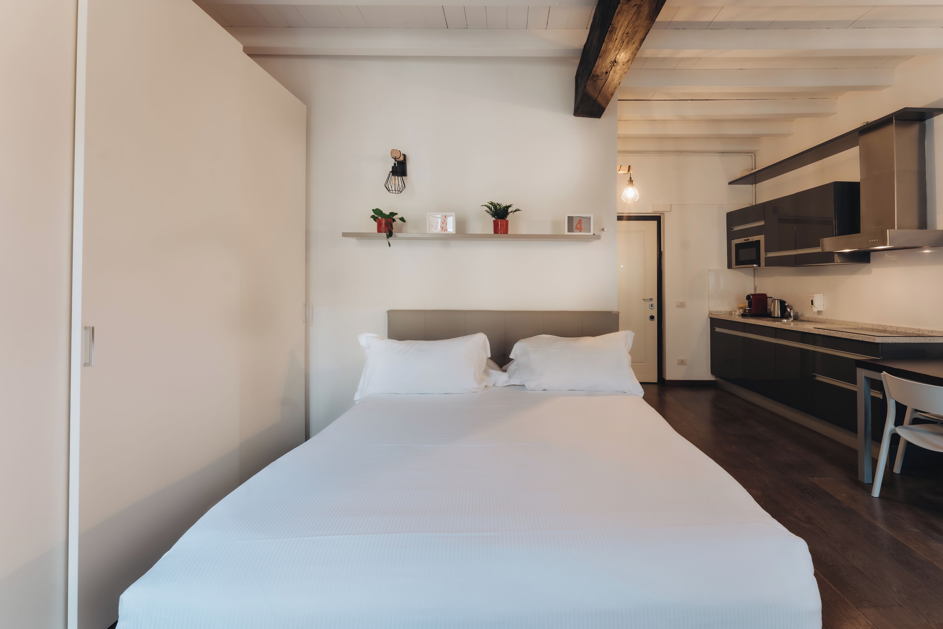 A modern hotel room with a large bed, a kitchenette, and a small shelf with decorative items.