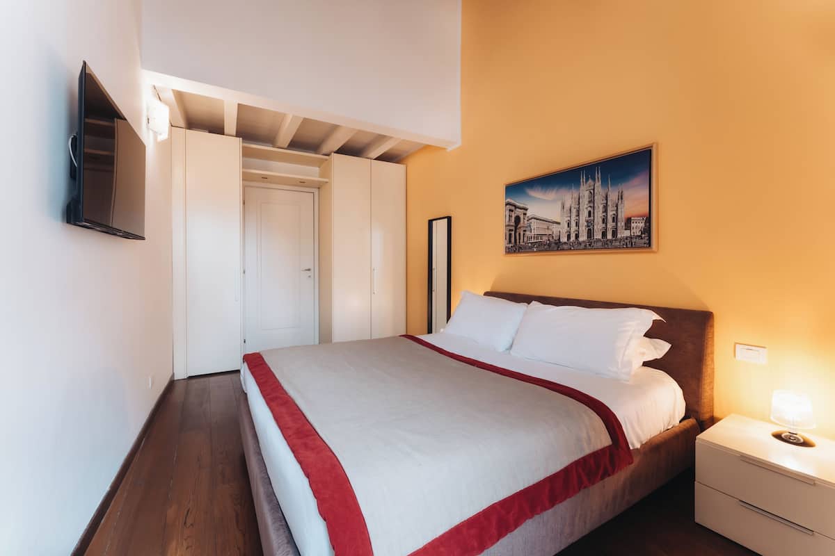 A neatly made bed with a red and white bedspread, a bedside lamp, and a framed picture on the wall.