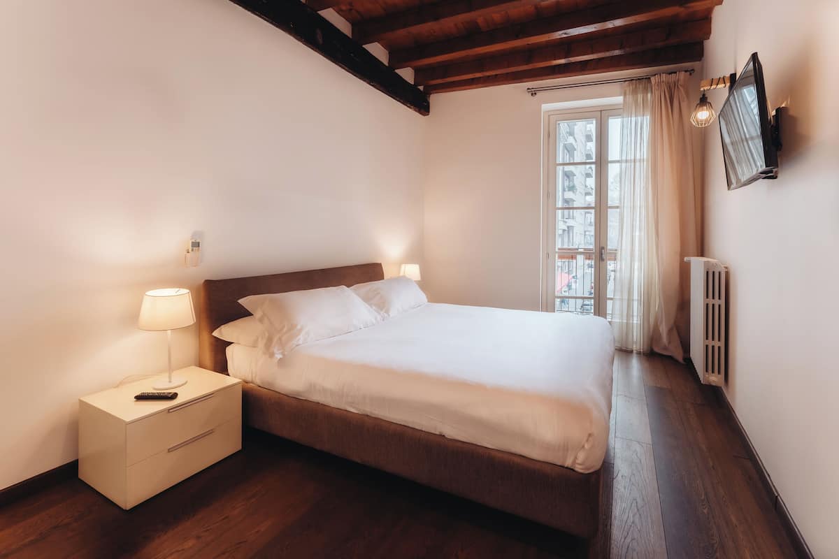 A bedroom with a wooden bed, a nightstand with a lamp, and a window with curtains.