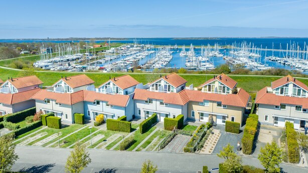 A marina with numerous sailboats, surrounded by residential buildings with red roofs and well-maintained gardens.