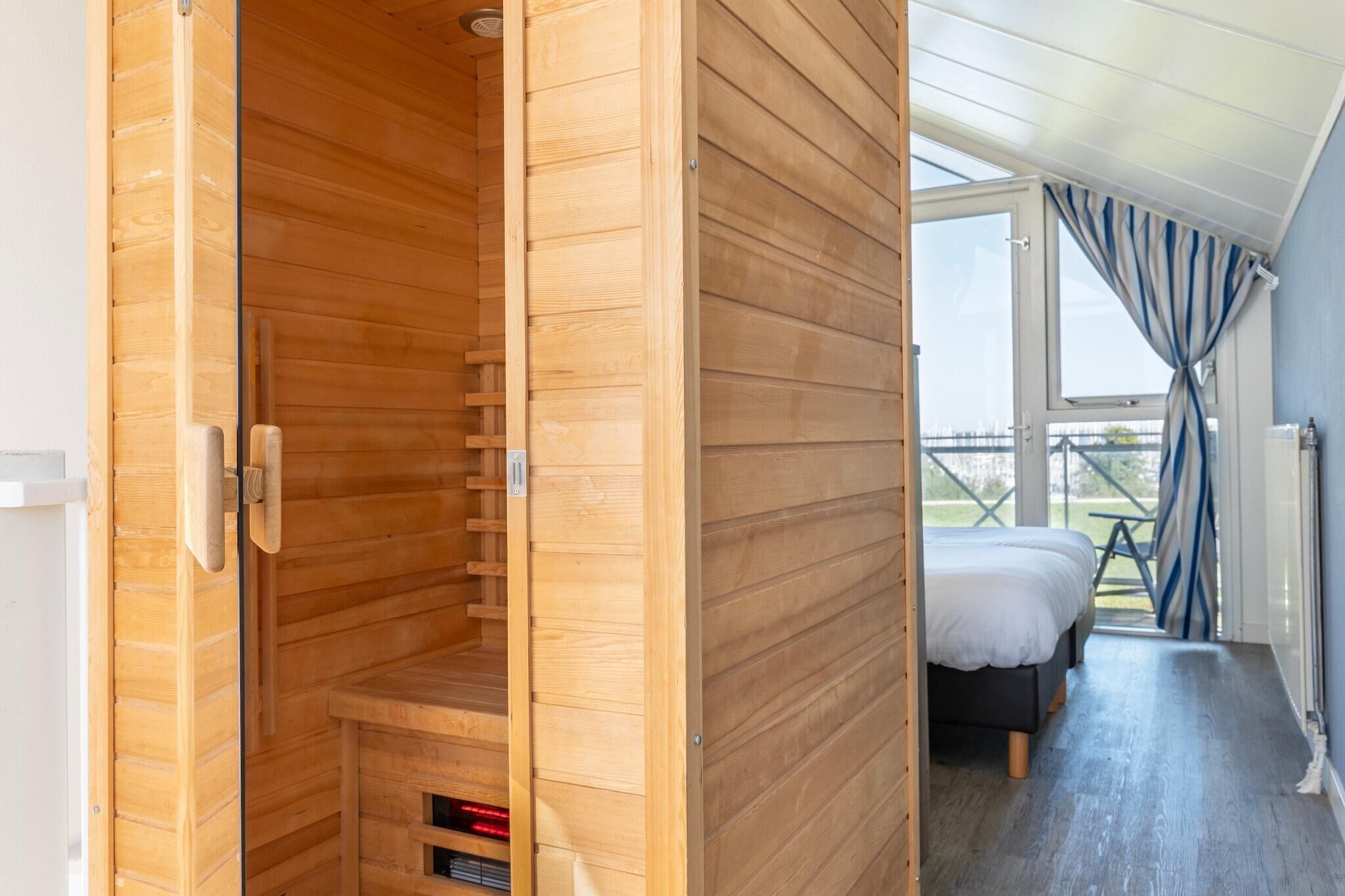 A wooden sauna room with a bench, a bed with white bedding, and a view of the outdoors through a large window.