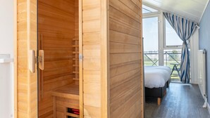 A wooden sauna room with a bench, a bed with white bedding, and a view of the outdoors through a large window.