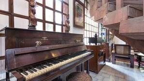 A grand piano in a spacious room with large windows, wooden beams, and a staircase. - Hotel Malhar Haveli (Noida)