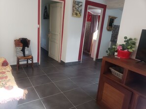 A living room with a sofa, a chair, a TV stand with a plant, and a hallway leading to a bathroom.