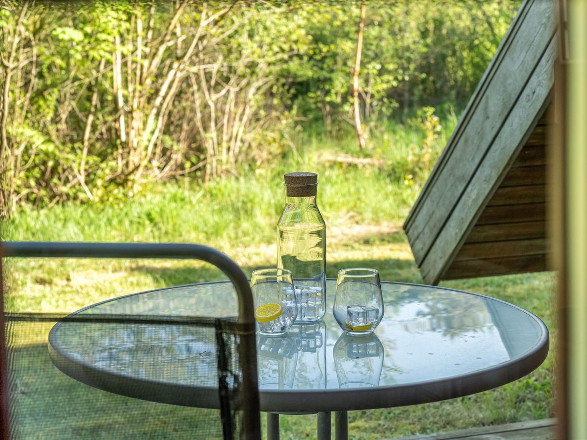 Een ronde glazen tafel met een fles en twee glazen erop, buiten geplaatst met uitzicht op groen en een houten constructie op de achtergrond.