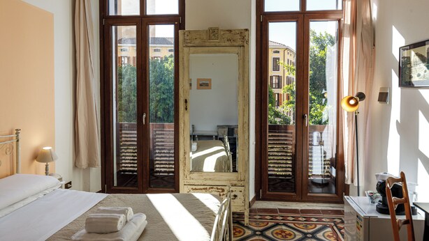 Ein Schlafzimmer mit einem Bett, einem großen Spiegel, einem Schreibtisch und Blick auf Gebäude durch die Fenster.