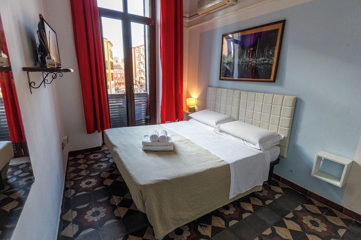 Une chambre d’hôtel avec un lit, une table de chevet, une fenêtre avec des rideaux rouges, une étagère fixée au mur et un tableau encadré.