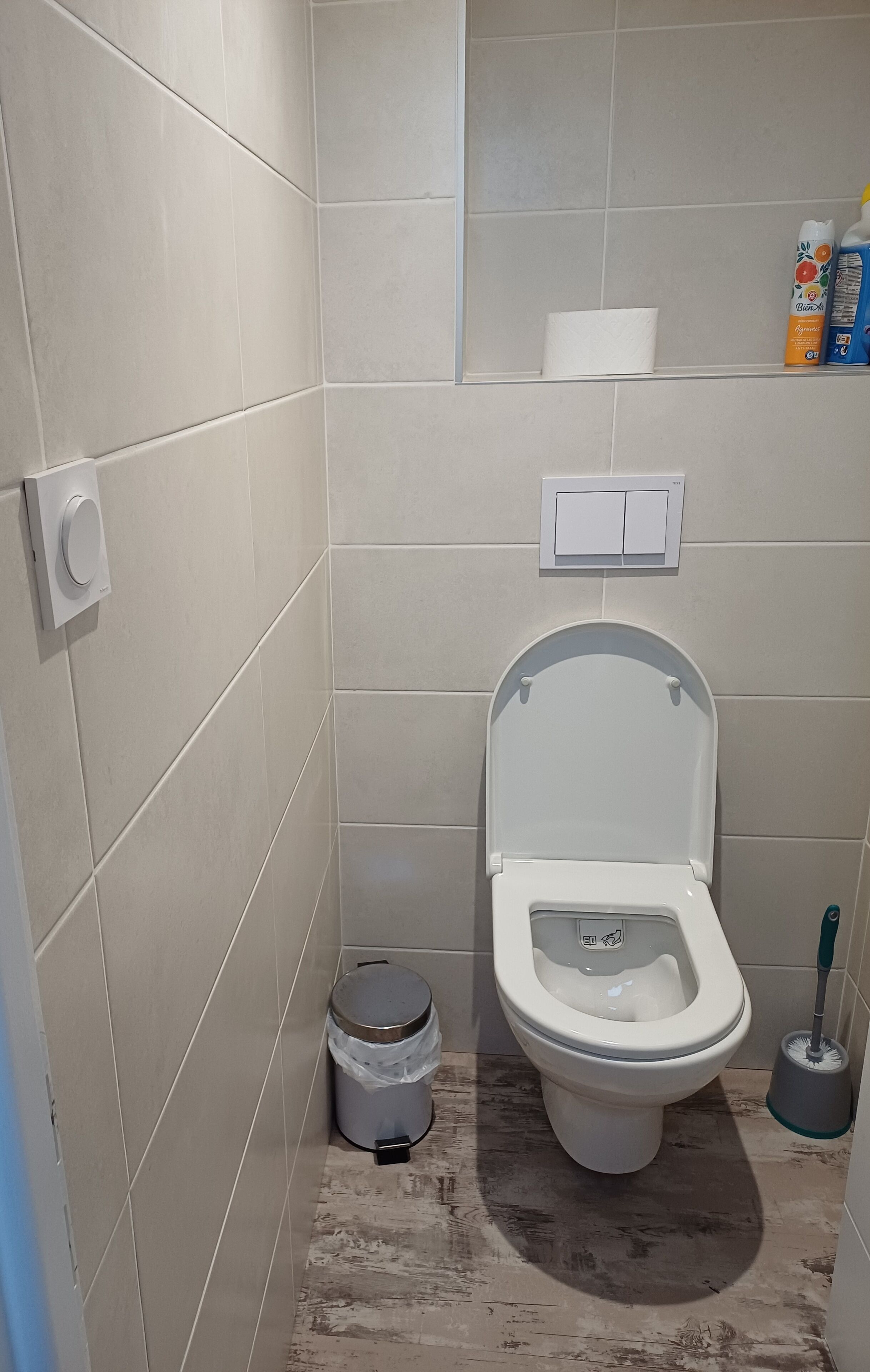 A small bathroom with a toilet, a trash can, a toilet brush, and a shelf with cleaning supplies.