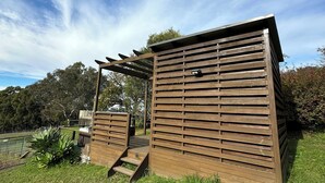 Una estructura de madera con techo de listones, una pequeña escalera y un área verde con árboles al fondo.