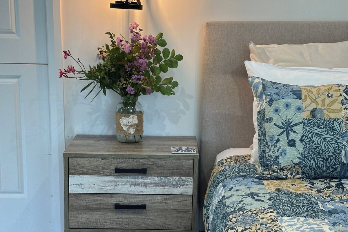 Un dormitorio con mesita de noche de madera, un florero con flores, ropa de cama floral y una lámpara colgante.