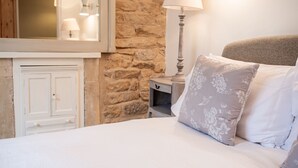 A bedroom with a stone wall, a white door, a bedside table with a lamp, and floral pillows. - Quaint and traditional dog friendly - Archway Cottage (Blockley)