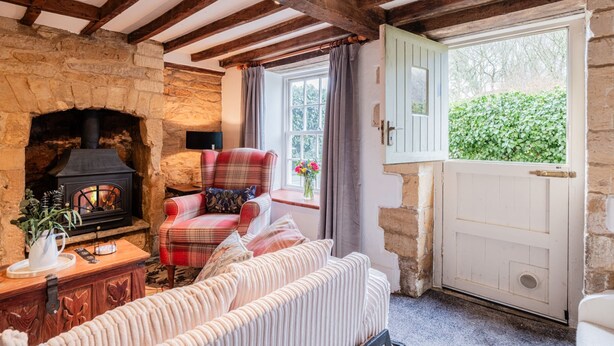 A cozy room with a stone fireplace, a plaid armchair, a cushioned bench, and a wooden door leading outside. - Quaint and traditional dog friendly - Archway Cottage (Blockley)