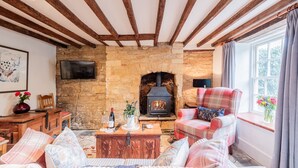 A cozy living room with exposed wooden beams, a stone fireplace, a plaid armchair, and a television mounted on the wall. - Quaint and traditional dog friendly - Archway Cottage (Blockley)