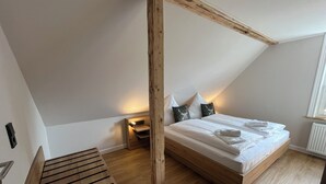 A bedroom with a sloped ceiling, a wooden bed frame, a bench, and a small bedside table with a lamp. - Über den Dächern von Braunlage - Urlaub zu Viert (Braunlage)