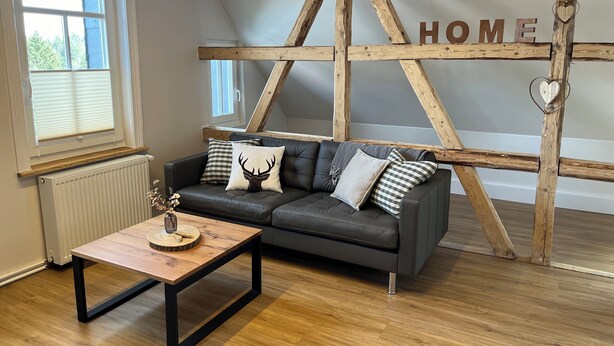 A cozy living room with a dark grey sofa, wooden beams, and a 'HOME' sign on the wall. - Über den Dächern von Braunlage - Urlaub zu Viert (Braunlage)
