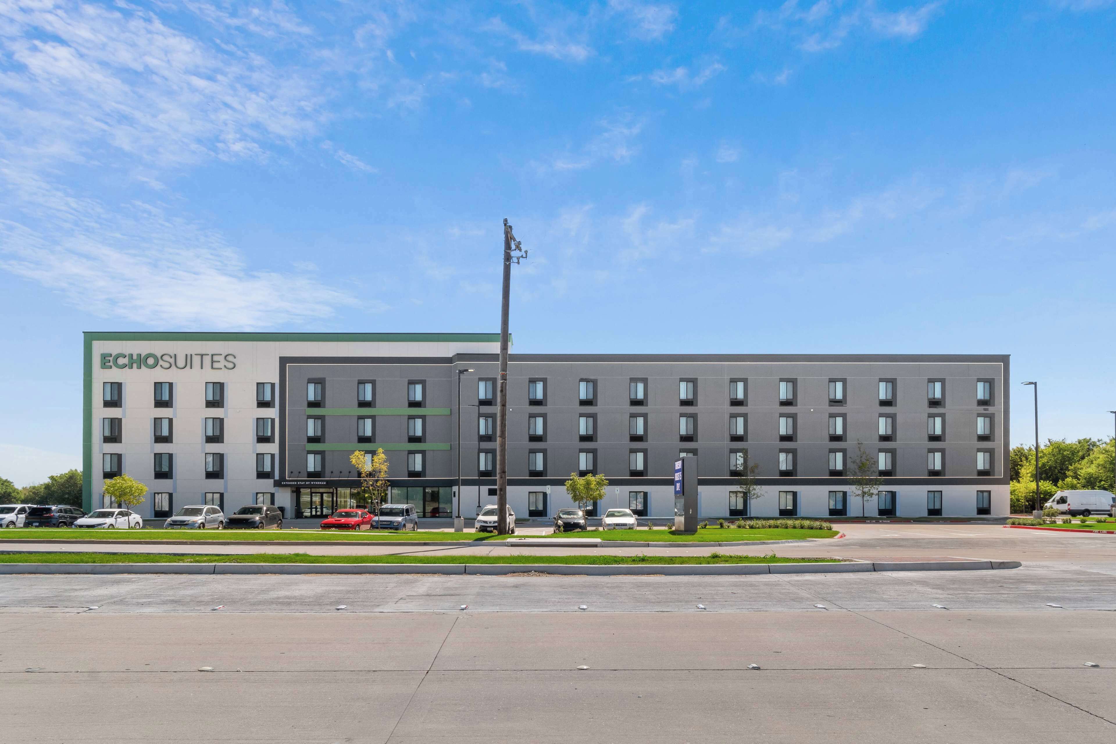 A modern hotel building with a flat roof, multiple windows, and a parking lot in front.