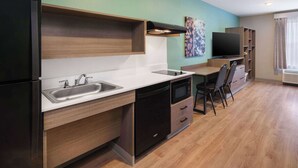 A compact kitchenette with a sink, microwave, and stove, a small dining area with a chair, and a TV on the wall.