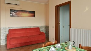 A room with a red sofa, a table with a green tablecloth, a painting on the wall, and a door with a light blue frame. - La Grolla del Garda - App. Giada (Manerba del Garda)