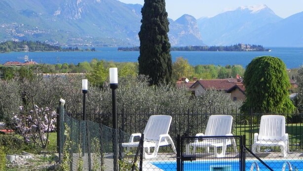 A pool area with white lounge chairs, a fenced pool, and a view of a lake with mountains in the background. - La Grolla del Garda - App. Giada (Manerba del Garda)