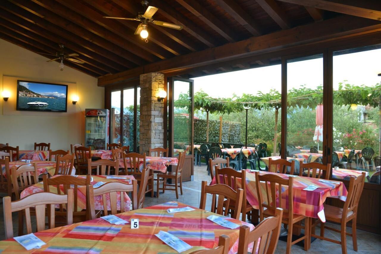 An outdoor restaurant with wooden tables and chairs, covered by a wooden roof, and large windows offering a view of the garden.