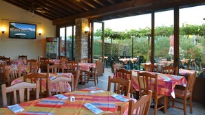An outdoor restaurant with wooden tables and chairs, covered by a wooden roof, and large windows offering a view of the garden. - La Grolla del Garda - App. Giada (Manerba del Garda)