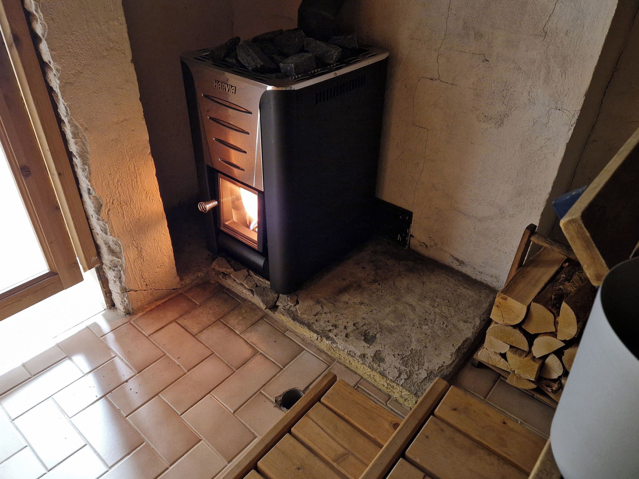 A wood-burning stove with a visible fire, a tiled floor, and a stack of firewood.